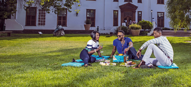 Three adults smile while having a picnic on a lush lawn in Bloemfontein, which can be experienced when you book your hotel or other accommodation with Flight Centre.